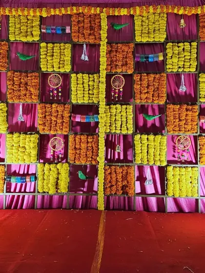 A colorful and traditional baby shower backdrop, featuring a grid of marigolds, pink drapes, and decorative elements like dreamcatchers and parrots.