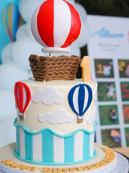 A charming hot air balloon cake for a first birthday celebration. The design features a red and white striped balloon topper and smaller balloons around the base, perfectly matching the party's adventurous theme.