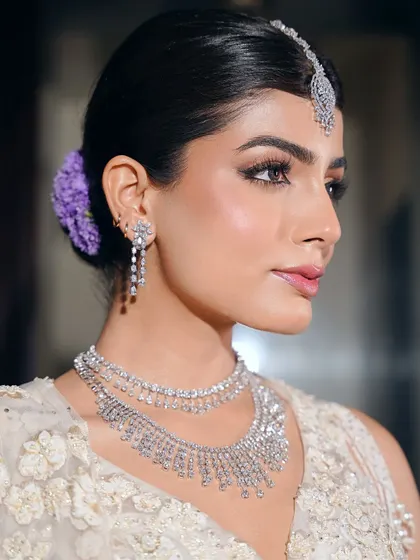 A close-up of the dewy bridal glam. The skin is luminous, the eyes are softly defined, and a hint of purple flowers in her hair adds a unique, modern touch to the classic white bridal look.