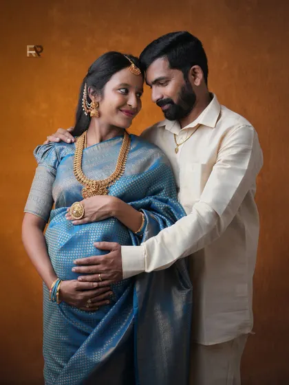 A loving embrace. The dad-to-be gently holds his partner, both dressed in beautiful traditional attire. The rich colors of the saree and the warm backdrop create a heartfelt image.