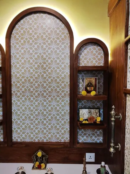 The interior of the Divinity Home pooja room, featuring arched wooden niches against a patterned wallpaper. The backlighting creates a warm, golden glow, enhancing the spiritual atmosphere.