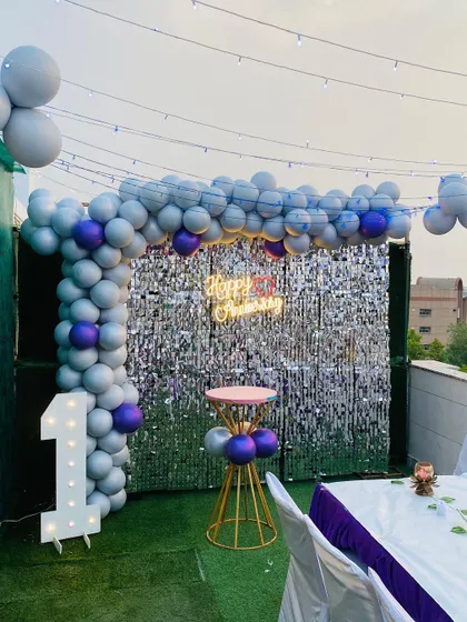 A stylish first birthday party on a rooftop terrace. The decor includes a balloon arch in grey and purple against a silver shimmer wall, a light-up marquee number, and string lights overhead for a festive evening glow.