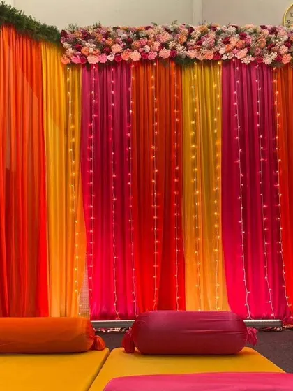 A beautiful backdrop of orange, pink, and yellow drapes with integrated fairy lights. This setup provides a warm and colorful background suitable for either a Mehendi or Sangeet event.