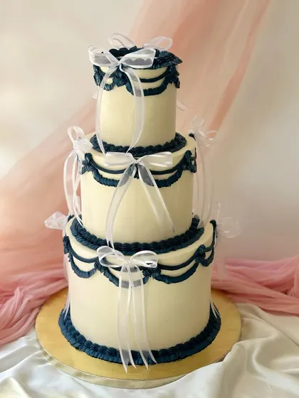 A striking three-tier cake featuring vintage-style navy blue Lambeth piping on a clean white buttercream base. Each tier is finished with delicate white ribbons for a classic, formal look.