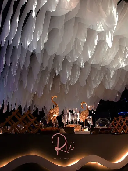 A modern bar setup with a unique, cloud-like ceiling installation made of white fabric. The golden flamingo figurines add a touch of playful luxury.