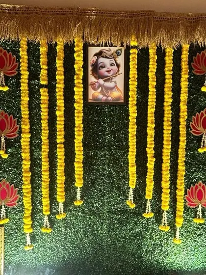 A Krishna-themed backdrop on a greenery wall. The design features hanging marigold garlands, lotus cutouts, and a central image of baby Krishna, ideal for naming ceremonies or Janmashtami.