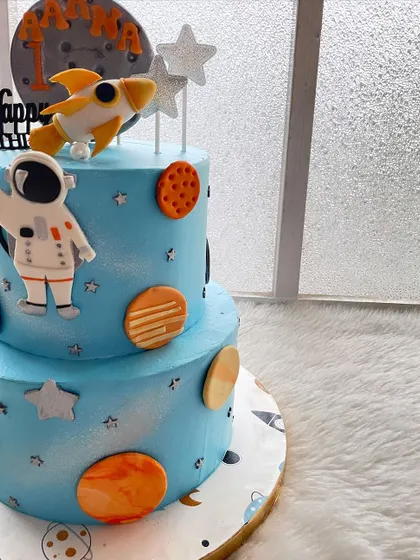 A two-tier space-themed cake for a first birthday. This design features a rocket, an astronaut, and lots of planets, all made with whipped cream and fondant toppers.