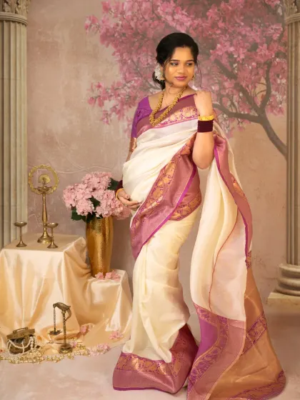 A full-length standing portrait of the mother-to-be, showcasing the beautiful drape and border of her cream and purple silk saree. The soft, painted backdrop adds a touch of romance to this traditional look.