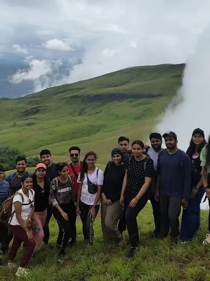 A corporate group poses on the grassy slopes of Netravati Peak, with a dramatic cloud-covered mountain in the background.