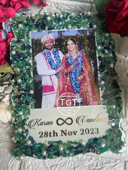 A 9x12 inch resin tabletop frame with a unique green and blue floral border made from a preserved varmala. The couple's names, infinity symbol, and wedding date are in gold.
