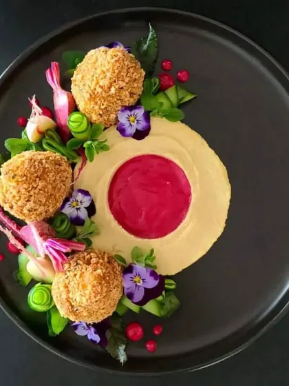 A beautifully plated appetizer of crispy croquettes on a bed of hummus and beet puree, garnished with edible flowers. This dish is as beautiful as it is delicious.