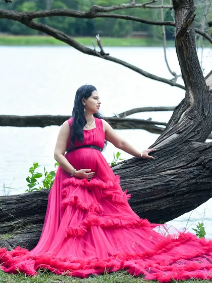 A striking portrait by the lake. The mom-to-be is seated on a fallen tree log, her vibrant pink gown cascading down, contrasting beautifully with the water and woods.
