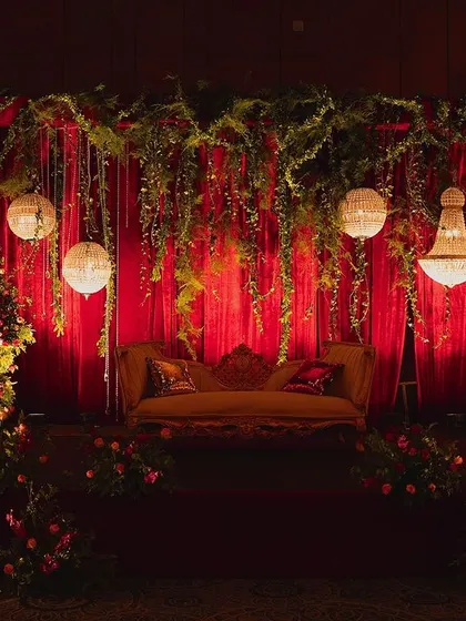 A stunning wedding stage where romance meets nature. We framed a classic couch with mountains of red and pink roses, hanging crystal globes, and a lush green canopy against a dramatic red backdrop.