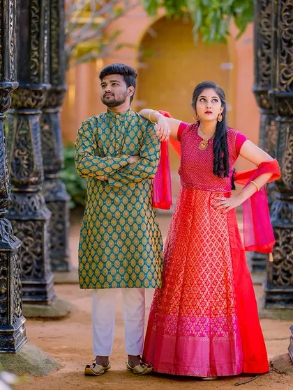 A stylish portrait of a couple in contrasting traditional wear. Their poses are confident and modern, set against a row of intricately carved pillars.