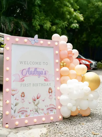 A close-up of the welcome sign for Aadhya's first birthday. The light-up frame and custom fairy-themed artwork, paired with a pastel balloon garland, create a warm and inviting entrance for guests.