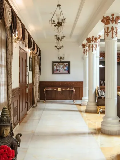 A long corridor in the palace, flanked by ornately carved wooden pillars on one side and stately white columns on the other, leading the eye towards a console at the far end.