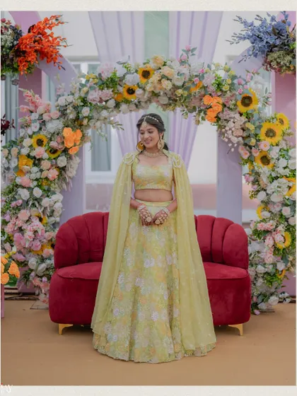 A full-length portrait of the bride in her pastel yellow Haldi lehenga, standing before a floral arch.