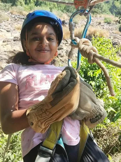 A young camper gets ready for her turn on the zipline, fully equipped with a helmet and gloves for safety.