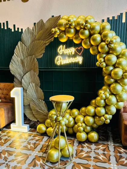 I designed this elegant and modern first-anniversary decoration in a restaurant. The setup features a full ring of metallic gold balloons, dried pampas grass accents, a 'Happy Anniversary' neon sign, and a lit marquee number '1'.