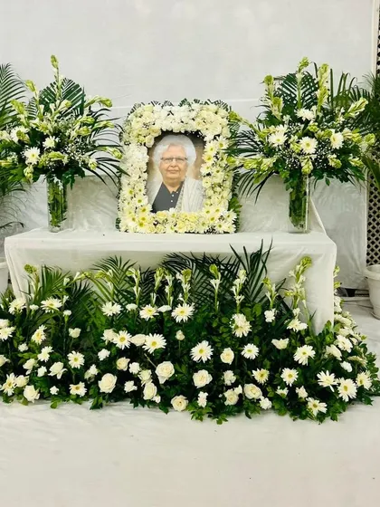 This is the kind of serene and respectful memorial setup I create for families. It features a central table with a beautiful photo frame garland made from fresh white flowers, flanked by elegant bouquets and lush green plants. I design these arrangements to bring a sense of peace and dignity to the service, ensuring every detail is handled with care.