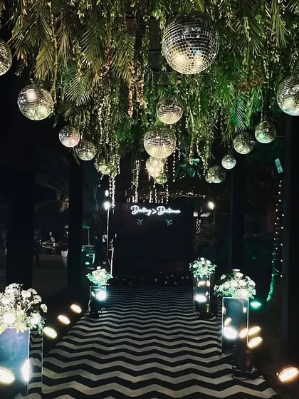 The entrance to a 'Dancing and Dining' themed sangeet. A chevron-patterned walkway leads guests under a ceiling of hanging disco balls and greenery, setting the stage for a night of fun.