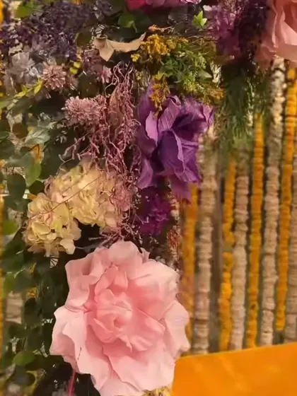 A close-up of the floral work in a Haldi decoration, showing a mix of pink and purple paper flowers against a backdrop of traditional marigold strings.