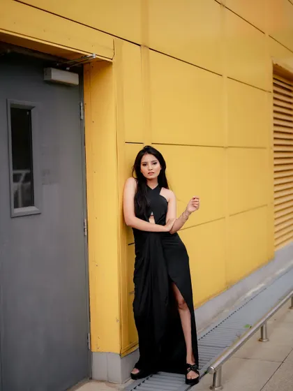 A full-body shot of a model in a black evening gown against a yellow industrial wall. The contrast between the elegant dress and the stark background creates a modern, high-fashion aesthetic.