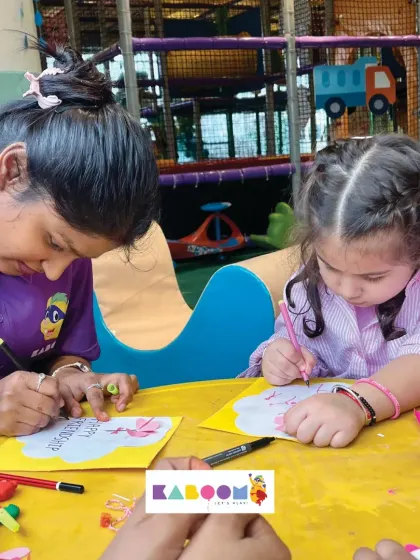 A staff member and a young girl sit side-by-side, both concentrating on drawing and coloring Friendship Day cards. Crafting together is a core part of our experience.