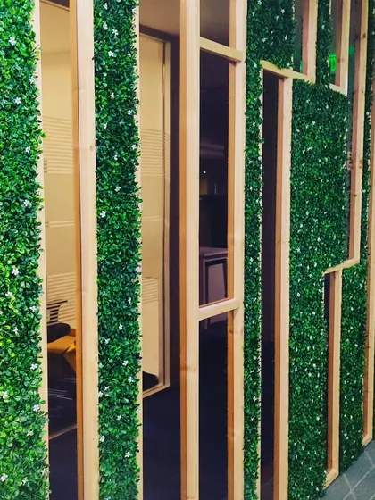 A custom office partition wall combining wood frames with vertical strips of artificial greenery. This design allows light to pass through while still providing a sense of separation.