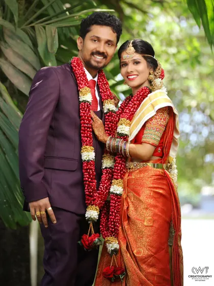 A happy couple portrait taken outdoors, with the groom in a stylish suit and the bride in a vibrant red saree.