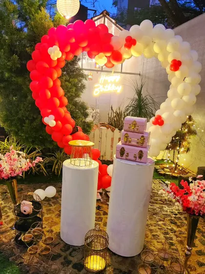 A heart-shaped balloon arch in red and white, set up in a garden for a romantic birthday or anniversary celebration.