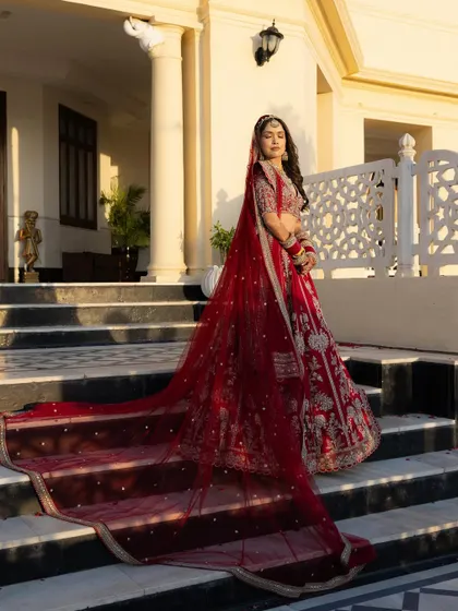A classic bridal portrait showcasing the bride, Sushmita, in her magnificent red lehenga. The long, flowing veil and regal pose against the palace architecture create a timeless and powerful image of bridal elegance.