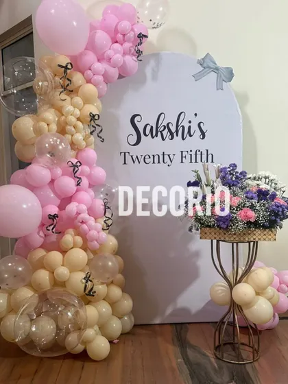 A beautiful pastel-themed setup for a 25th birthday. This decoration features a custom arched backdrop personalized with the name 'Sakshi', framed by a delicate balloon arrangement in shades of pink and nude.