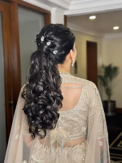 A glamorous and curly ponytail for Damini's reception. This hairstyle is adorned with delicate crystal accessories, adding a touch of sparkle to the modern and chic look.