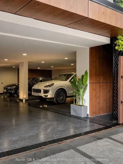 The entrance to the Meghana Foods corporate office, where dark brown gates with muted grooves slide open to a parking lot tiled in leather-finish granite. Potted plants and alternating glass and metal sheets create a sophisticated transitional space.