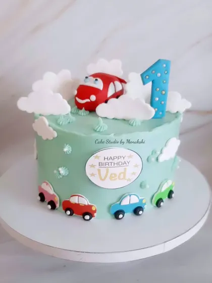 A gorgeous car themed first birthday cake. This design features a cute red car on top, surrounded by fondant clouds and smaller cars around the base.
