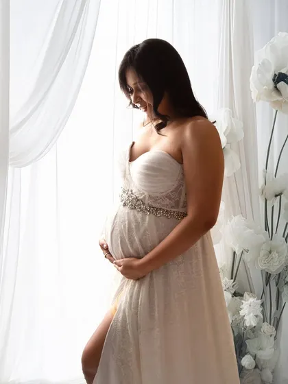 A classic window-lit portrait. The mom-to-be looks down at her bump, her white lace gown glowing in the soft light.