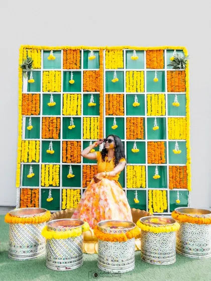 The bride strikes a pose in front of her vibrant Haldi decor. The colorful backdrop of marigolds and the traditional setup perfectly frame this fun and stylish portrait.