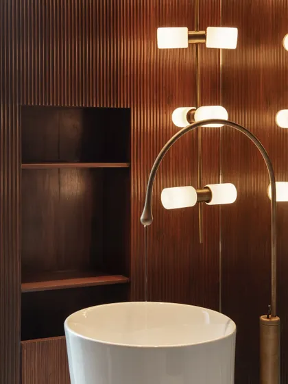 A powder room detail showing a custom-designed brass faucet and sculptural light fixtures against a fluted wood wall, demonstrating our focus on craft and materiality.
