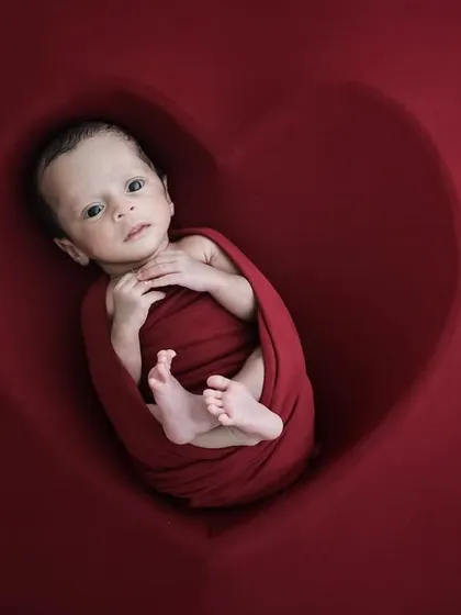 An awake baby wrapped in red, looking at the camera from inside our heart-shaped posing prop.