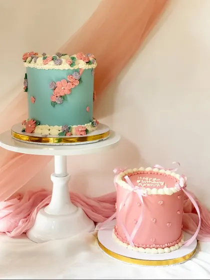 A pair of complementary vintage-style cakes. One is a blue floral wreath cake, and the other is a pink cake with delicate piping and ribbon details.
