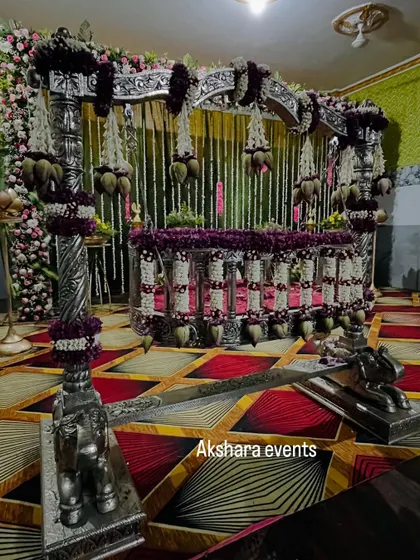 A grand silver cradle, intricately decorated with purple and white flowers, ready for a naming ceremony. The backdrop features hanging jasmine garlands for a classic touch.