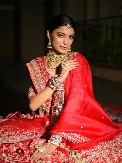 A full-length seated portrait, showing the grandeur of the red lehenga. The makeup is balanced and elegant, perfect for this regal look.