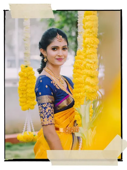 An artistic portrait of a bride in a vibrant yellow and blue saree, framed by hanging marigold flowers.