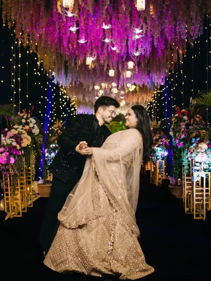 The couple shares a dance, lost in each other's eyes beneath a stunning canopy of purple wisteria and warm lights. This photo captures the romance and elegance of a beautifully decorated engagement night.