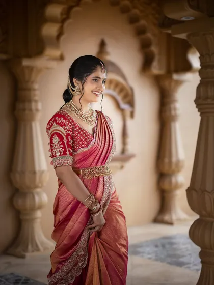 A candid portrait of the bride in a beautiful red saree, her smile captured in a moment of happiness. The architectural details of the venue add a touch of grandeur.
