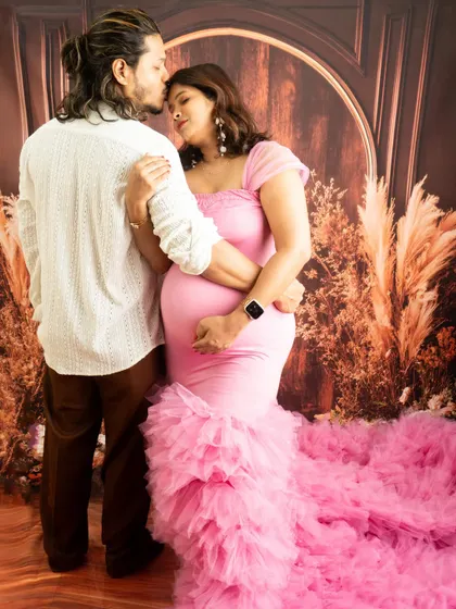 An intimate embrace from behind, showcasing the baby bump. The matching pink and white outfits create a soft and romantic color palette.