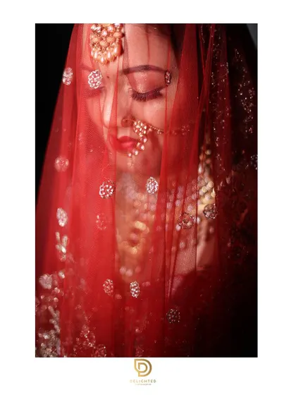 A classic bridal portrait with the red veil or 'ghunghat' drawn over her face. The soft light filtering through the fabric creates a delicate and timeless look.