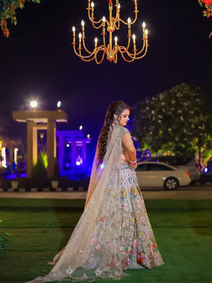 A magnificent full-length portrait of the bride in her multi-colored floral lehenga. The outdoor evening setting with a grand chandelier creates a fairytale-like scene.