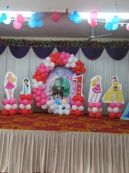 A grand 3D Princess theme stage decoration for a first birthday party. The setup includes multiple princess cutouts like Barbie, Jasmine, and Elsa, a balloon arch, and a personalized photo backdrop.